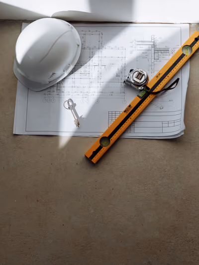 Overhead shot of an architectural house floor plan with room dimensions marked, laid on a table topped with a white hard hat, a house key, a spirit level, and a tape measure