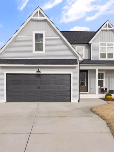 Completed two-story farmhouse-style home ready for move-in, with light gray vinyl siding, dark double-gable roofline, covered front porch, paved driveway, and a landscaped, mowed front lawn
