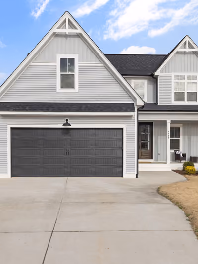 Completed two-story farmhouse-style home ready for move-in, with light gray vinyl siding, dark double-gable roofline, covered front porch, paved driveway, and a landscaped, mowed front lawn