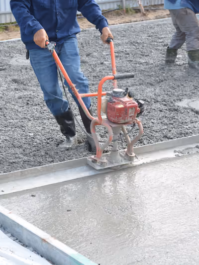 Wet concrete slab foundation being screeded and power-troweled by a construction worker, with a stacked gravel base, rebar mesh, and metal edge forms visible around the freshly poured section