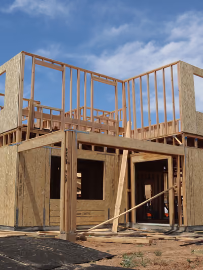 Two-story house under construction showing complete wood-frame skeleton with OSB-sheathed exterior walls, open stud window and door cutouts, and lumber scraps on the dirt lot