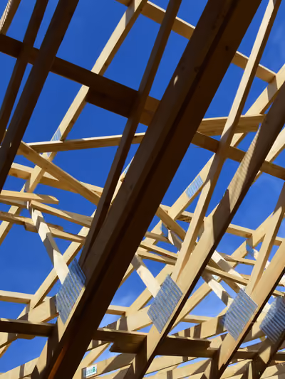 Close-up, low-angle view of exposed wooden roof trusses under construction, with visible galvanized metal gang-plate connectors joining the rafters and open sky showing through the unsheathed roof