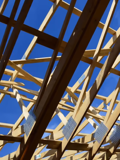 Close-up, low-angle view of exposed wooden roof trusses under construction, with visible galvanized metal gang-plate connectors joining the rafters and open sky showing through the unsheathed roof