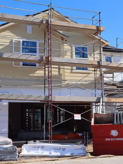 Multi-level pipe scaffolding erected across the full facade of a two-story home with pale yellow siding and finished windows, part of an active new-construction neighborhood