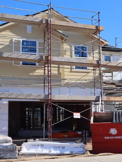 Multi-level pipe scaffolding erected across the full facade of a two-story home with pale yellow siding and finished windows, part of an active new-construction neighborhood