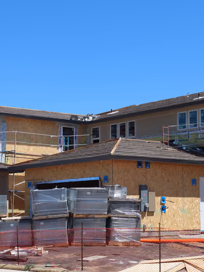 Two-story home mid-construction where one wing already has finished tan siding and windows while the other wing is still bare OSB wall sheathing wrapped in house wrap, with scaffolding and stacked window pallets on-site