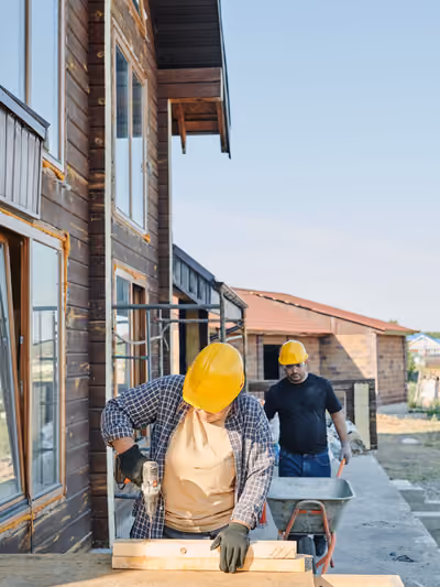 Two builders in yellow hard hats on an active residential job site: one drilling into a wood beam in the foreground, the other pushing a wheelbarrow past a house with unfinished wood siding and window openings