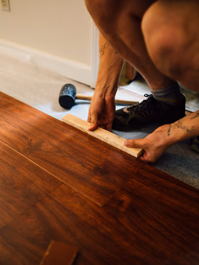 Installer using a rubber mallet and tapping block to fit a dark walnut-stained wood plank into place during flooring installation