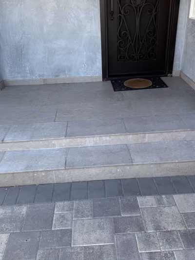 Newly renovated covered front entryway with large-format gray tile steps, a dark wrought-iron security door, and a matching paver walkway