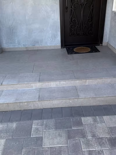 Newly renovated covered front entryway with large-format gray tile steps, a dark wrought-iron security door, and a matching paver walkway