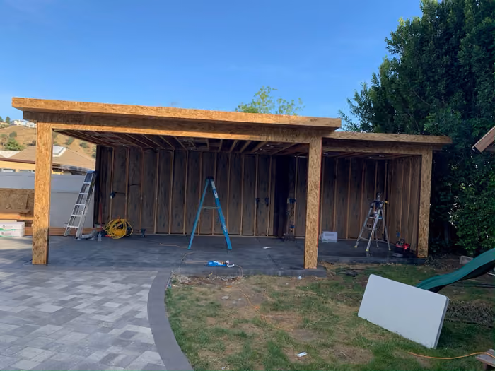 The covered patio mid-build with exposed wood framing, ladders, and tools on a bare concrete slab