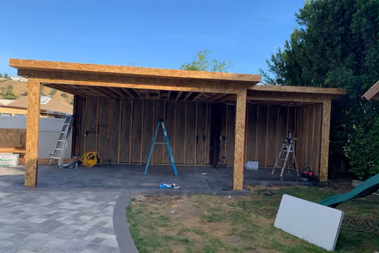 Backyard patio cover under construction, showing exposed wood post-and-beam framing on a new concrete slab