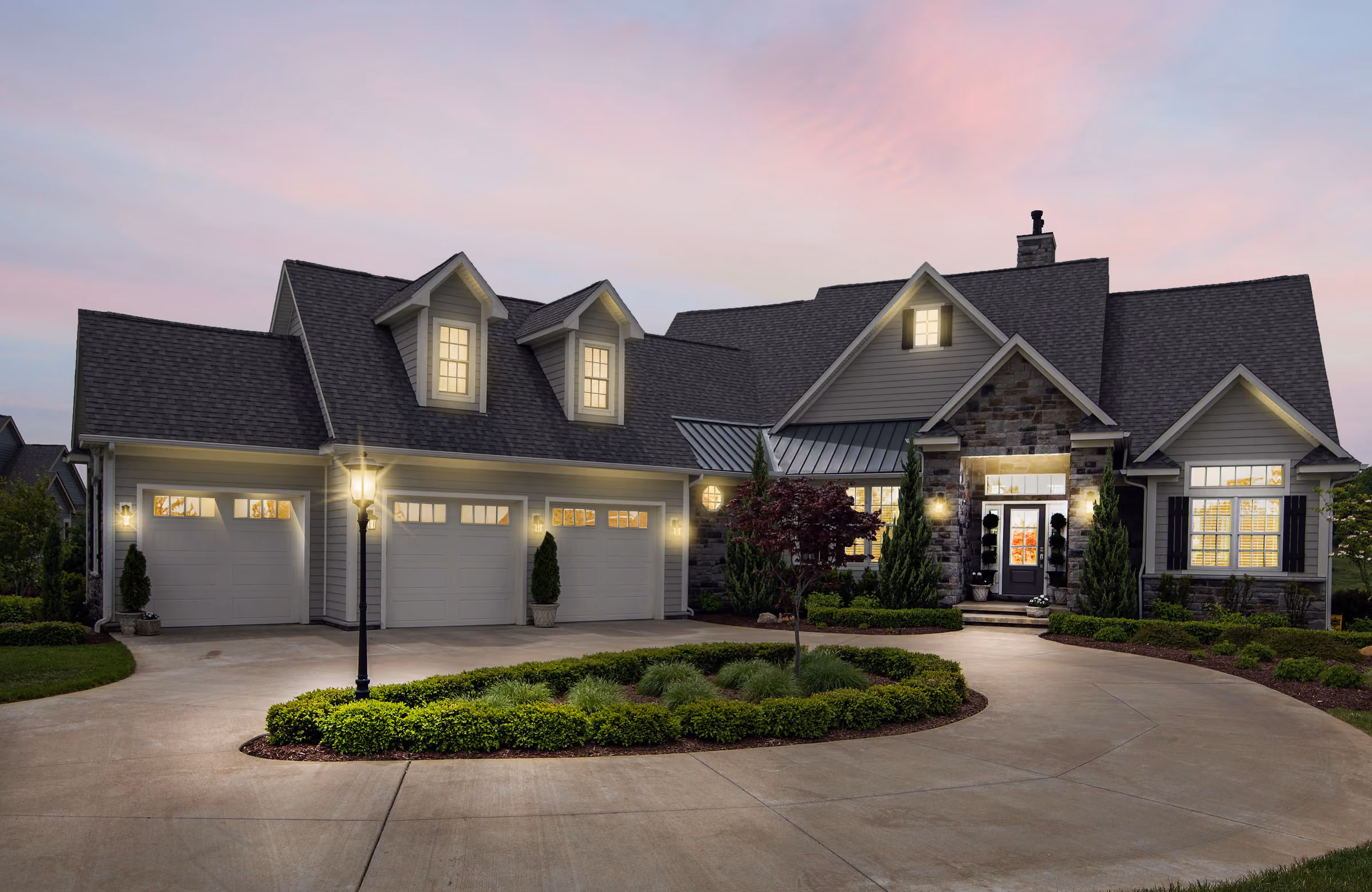 Custom-built home at dusk with warmly lit windows, a stone and shingle facade, and a landscaped circular driveway