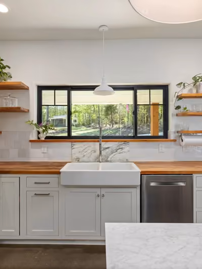 Farmhouse-style kitchen with white shaker cabinets, an apron-front farmhouse sink, butcher-block countertops, subway tile backsplash, and open wood shelving displaying dishware