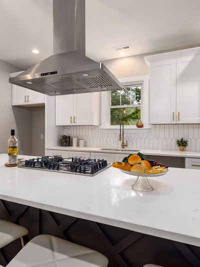White shaker-cabinet kitchen with a large center island featuring a dark X-braced base, white quartz countertops, stainless gas cooktop, and brass hardware