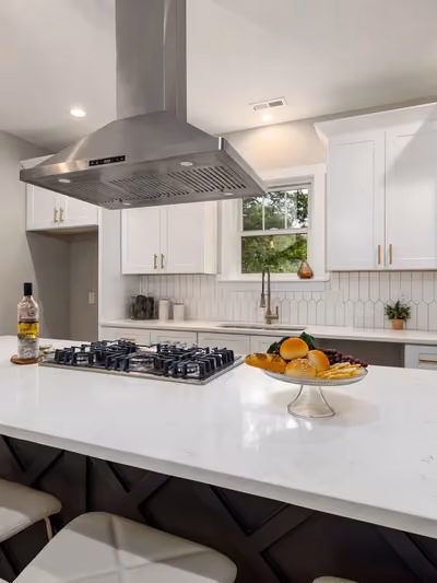 White shaker-cabinet kitchen with a large center island featuring a dark X-braced base, white quartz countertops, stainless gas cooktop, and brass hardware