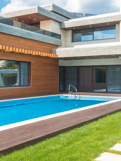 Modern luxury home exterior with wood-and-stone facade, a rectangular pool, wood deck, and manicured lawn