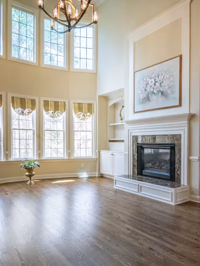 Two-story great room with a wall of tall arched windows, a marble fireplace flanked by built-in cabinetry, and rich hardwood flooring