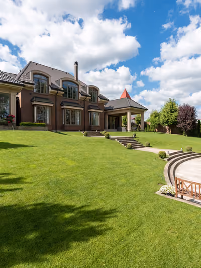Elegant two-story brick mansion exterior with a turret, arched dormer windows, and a manicured lawn with a curved paver walkway