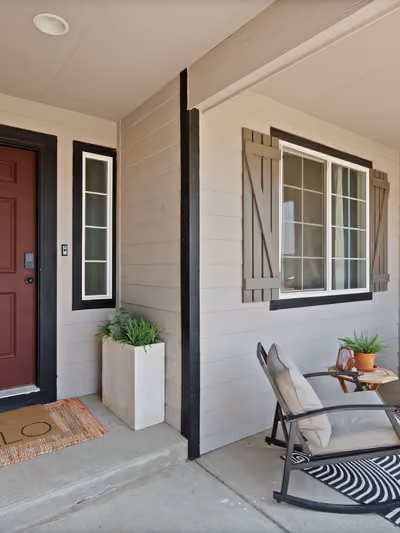 Covered front porch entryway with tan horizontal siding, black window trim and board-and-batten shutters, a maroon front door, and black metal rocking chairs