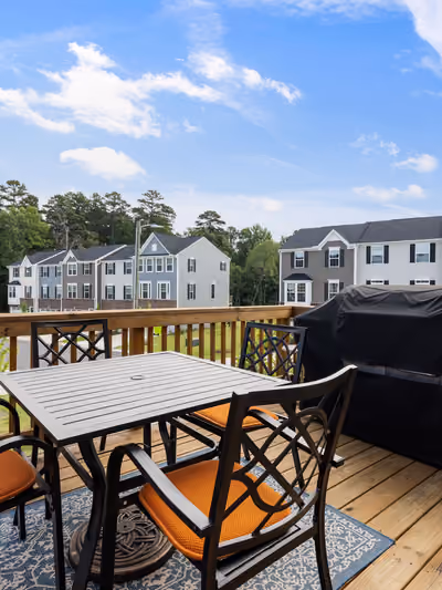 Elevated backyard wood deck with a metal dining table, four cushioned chairs, a covered gas grill, and a neighborhood view beyond the railing