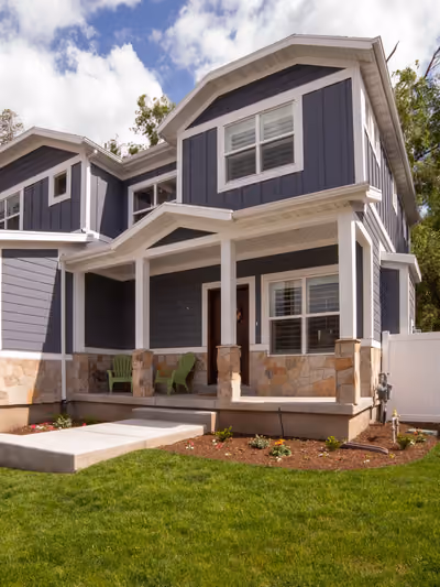 Two-story home exterior with dark blue-gray board-and-batten siding, white trim, a covered front porch with stone-clad columns, and mountain views in the background