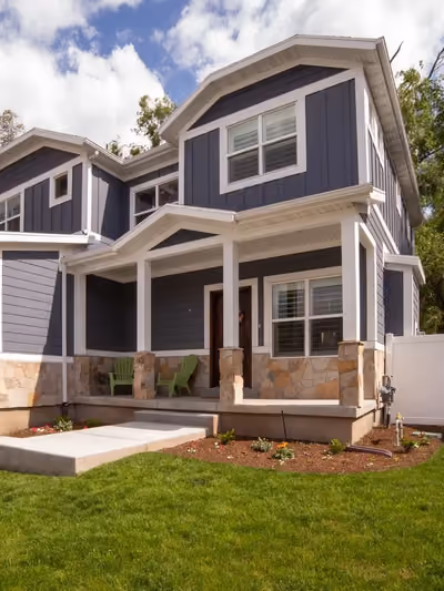 Two-story home exterior with dark blue-gray board-and-batten siding, white trim, a covered front porch with stone-clad columns, and mountain views in the background