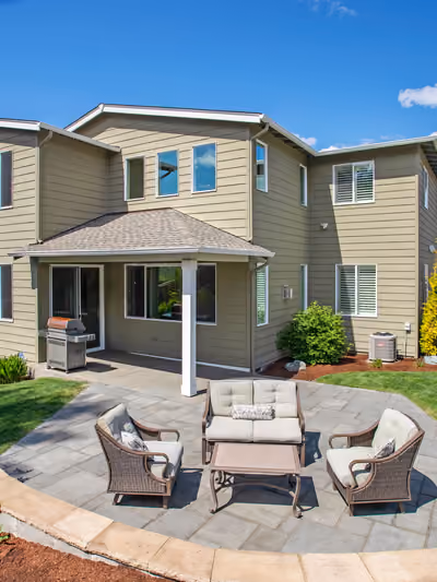 Landscaped backyard with a curved gray paver patio bordered by a stone edging wall, mulched planting beds, ornamental shrubs, and a cushioned wicker seating set