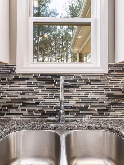 Close-up of a kitchen backsplash with dark and beige linear glass-mosaic tile, framed by white cabinets, a window, and a granite countertop with stainless double sink