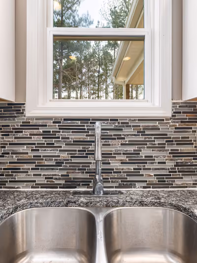 Close-up of a kitchen backsplash with dark and beige linear glass-mosaic tile, framed by white cabinets, a window, and a granite countertop with stainless double sink